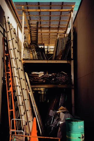 Tall ladders and stacked materials crowd a covered storage passage beside a green barrel.