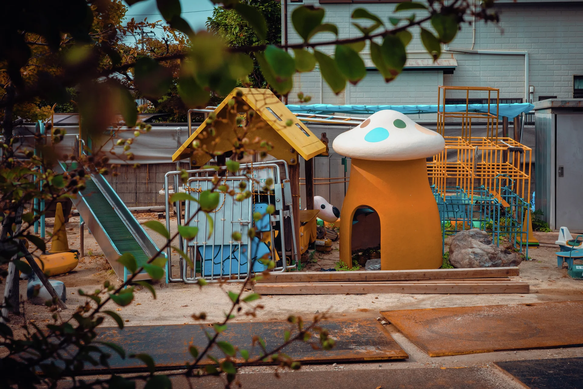 A small playground contains a slide, climbing frames, and a yellow mushroom-shaped structure behind leafy branches.