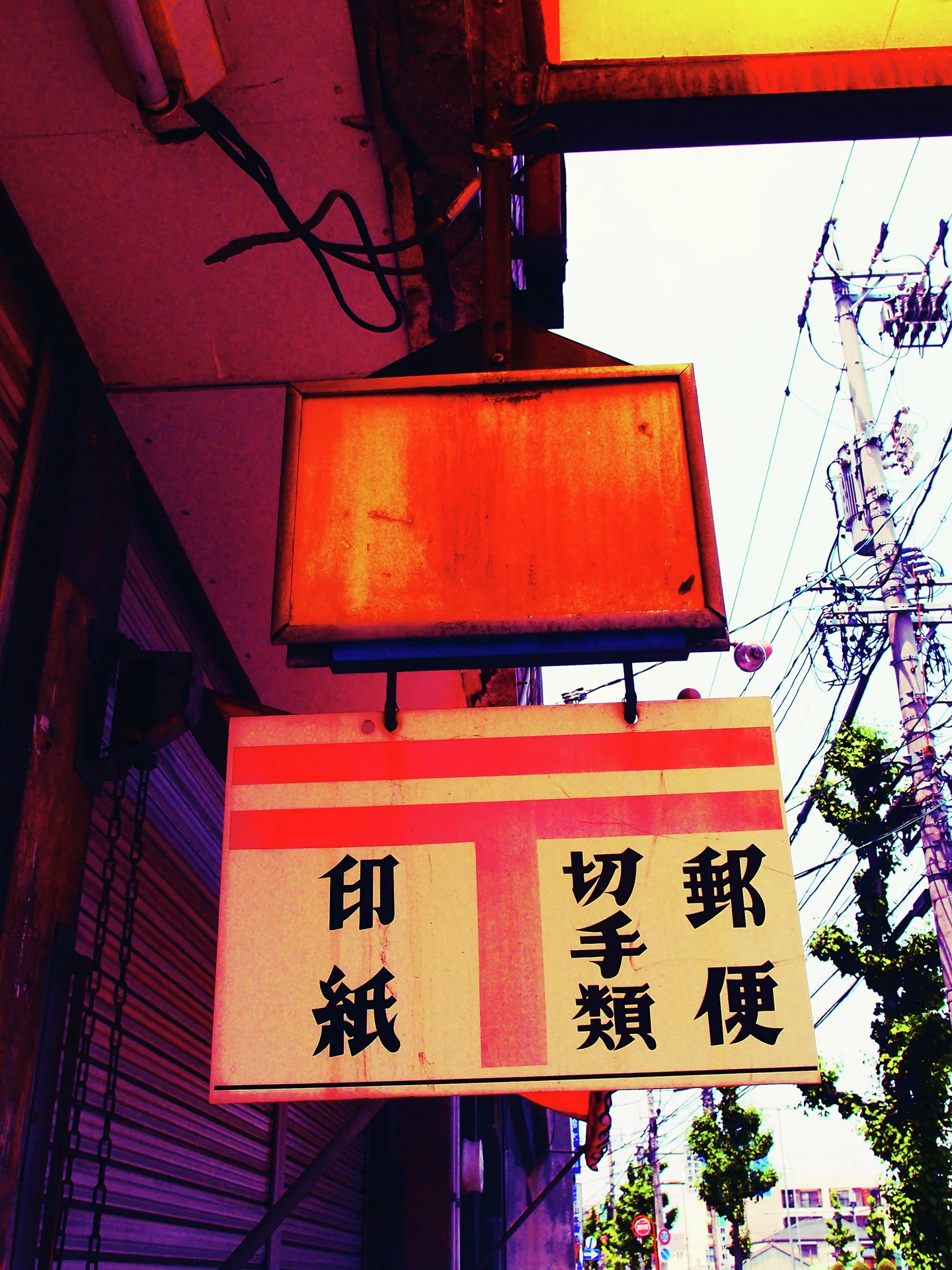 Two rectangular hanging signs project over a shuttered storefront beside utility wires and a leafy tree.