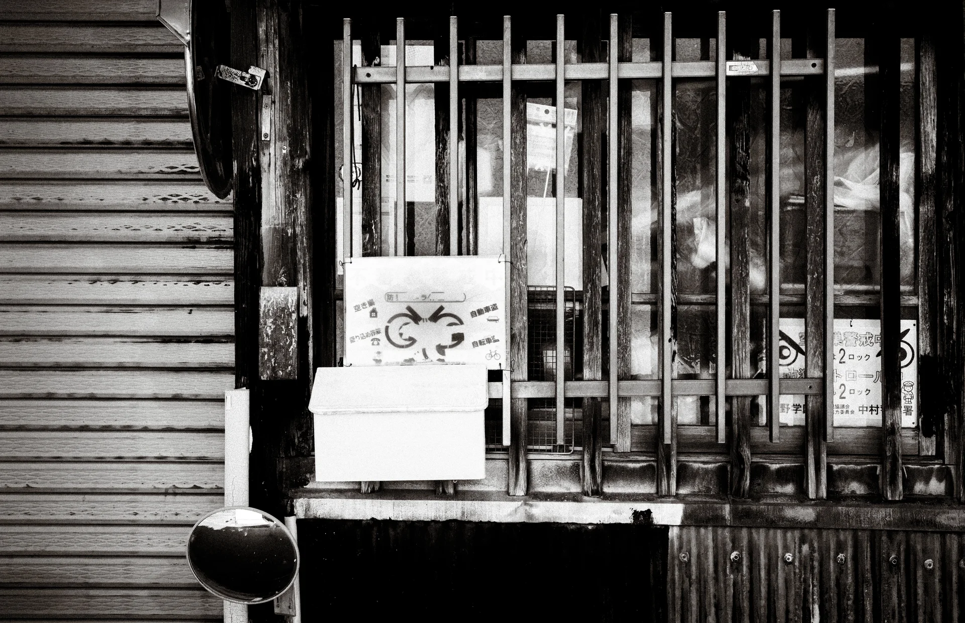 A barred wooden window holds notices and a small box beside a closed shutter in black and white.