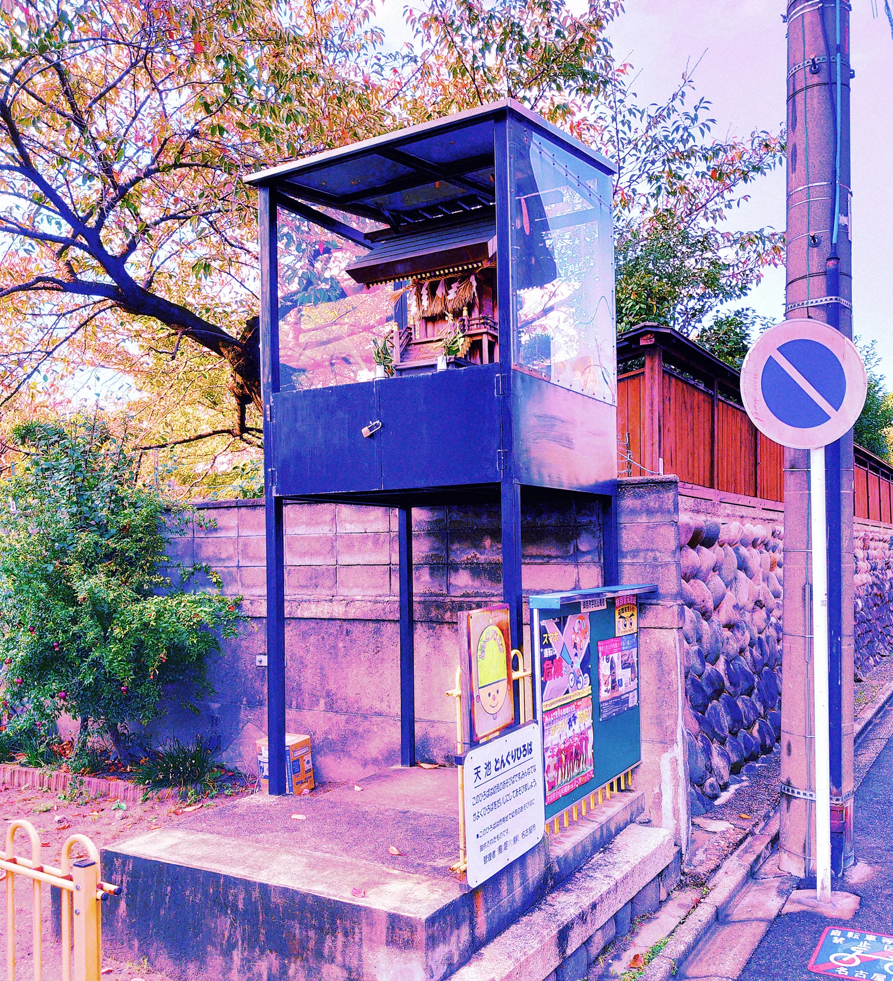 A blue open-sided metal structure stands above a stone base beside trees and a circular street sign.