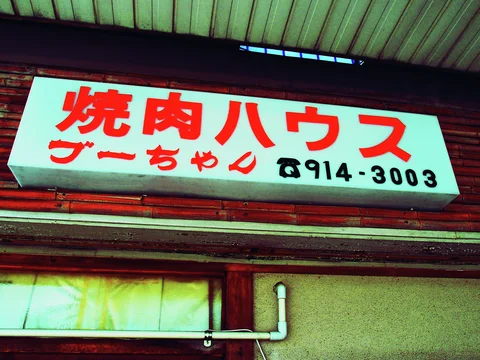 Bold red Japanese characters and a black phone number cover a pale projecting sign above weathered wood.