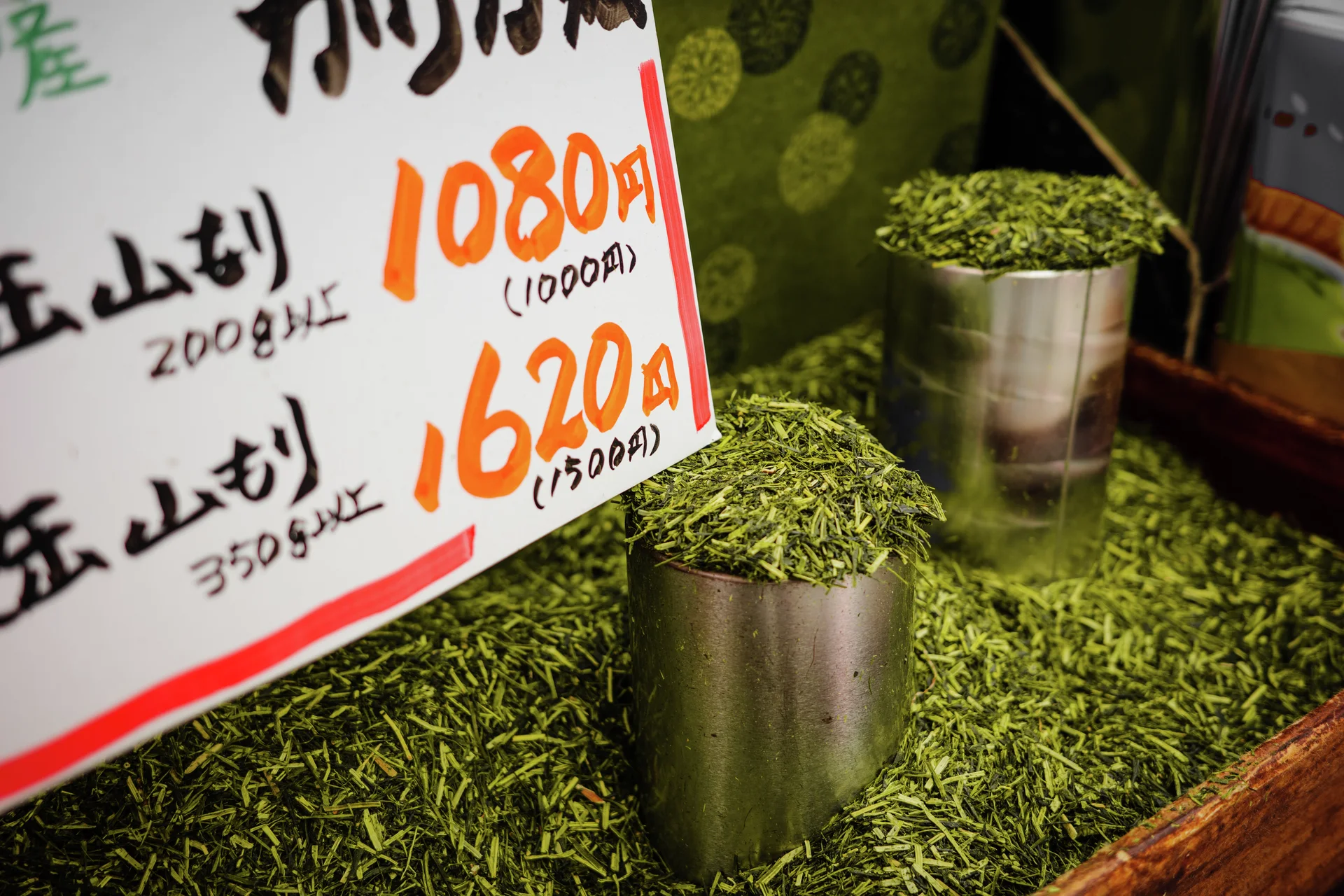 Metal measuring cups stand amid loose green tea leaves beside a handwritten price sign.