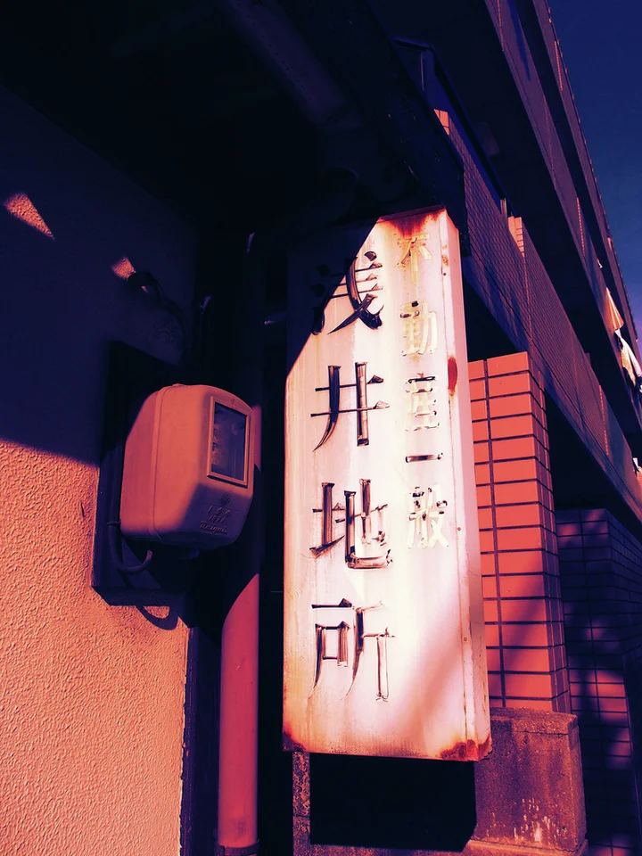 A pale rust-edged projecting sign bears dark vertical characters beside a brick wall and air-conditioning unit.
