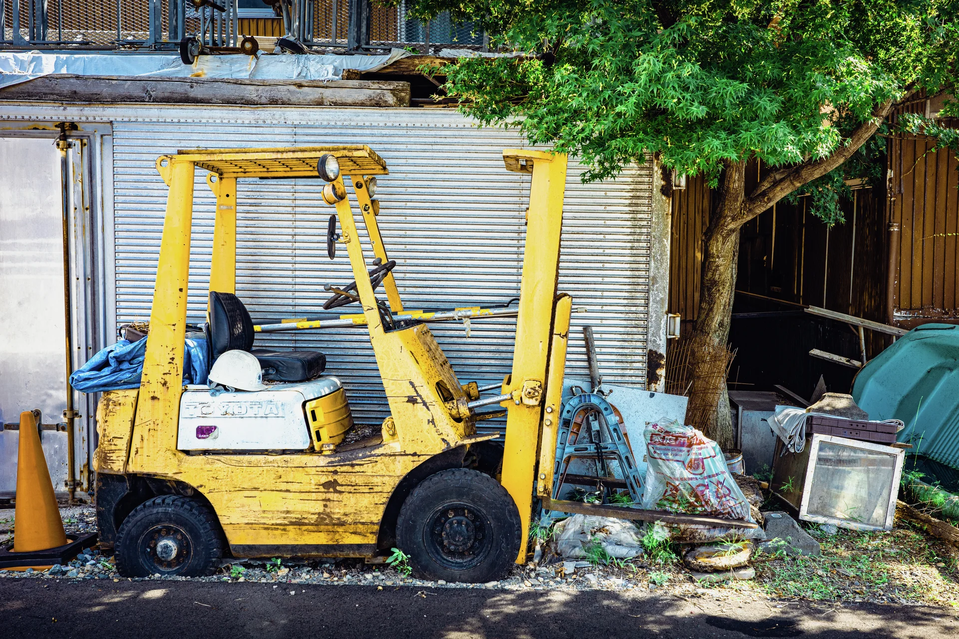 A weathered yellow forklift stands beside a tree amid scattered frames and equipment outside closed shutters.