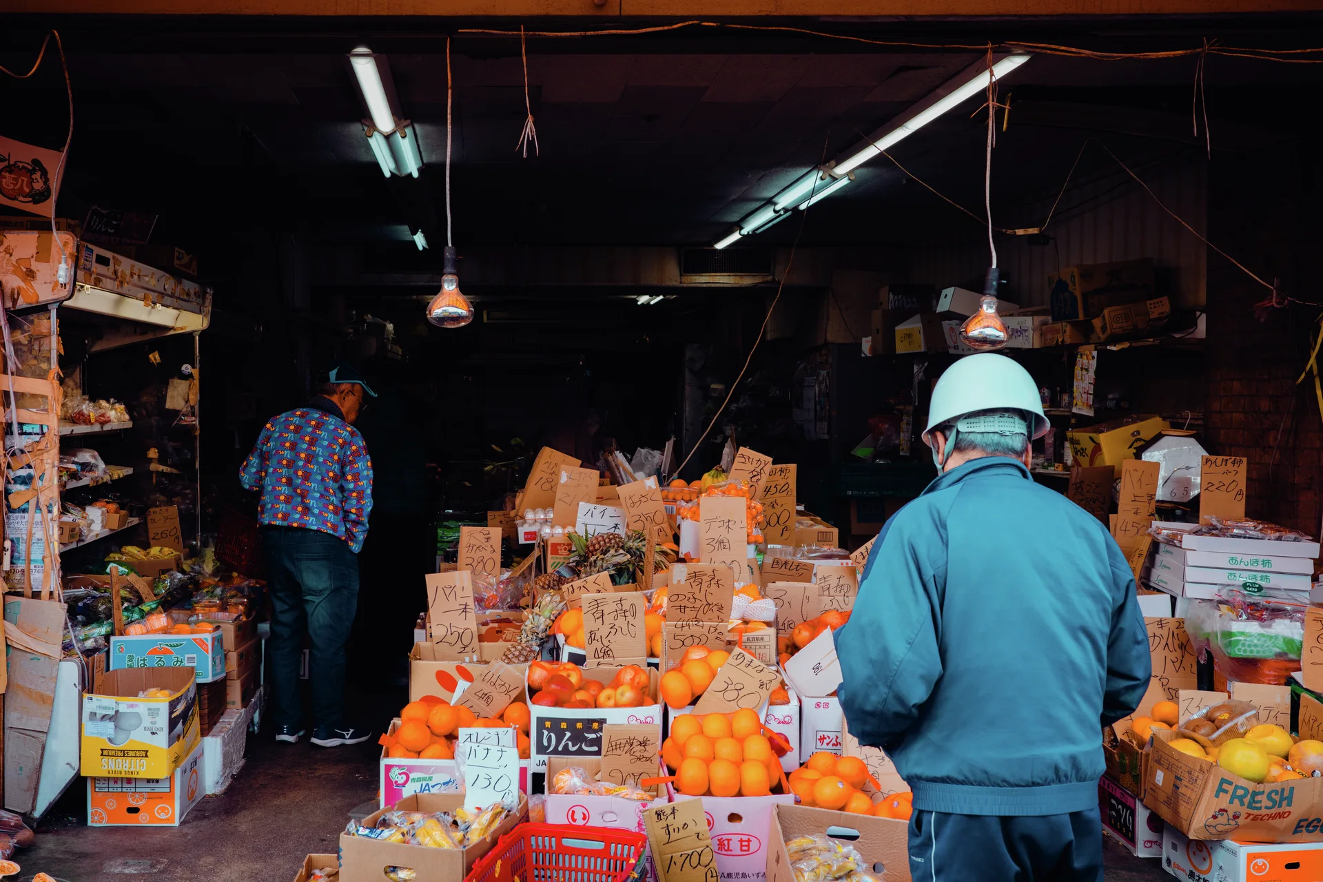 Two people stand inside a produce shop surrounded by boxes of oranges beneath fluorescent lights.