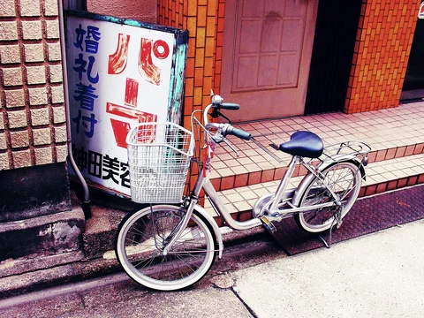 A silver bicycle with a wire basket stands beside a faded vertical salon sign and tiled entrance steps.
