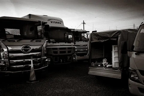 Several large trucks crowd a gravel yard beside an open tarpaulin-covered vehicle in black and white.