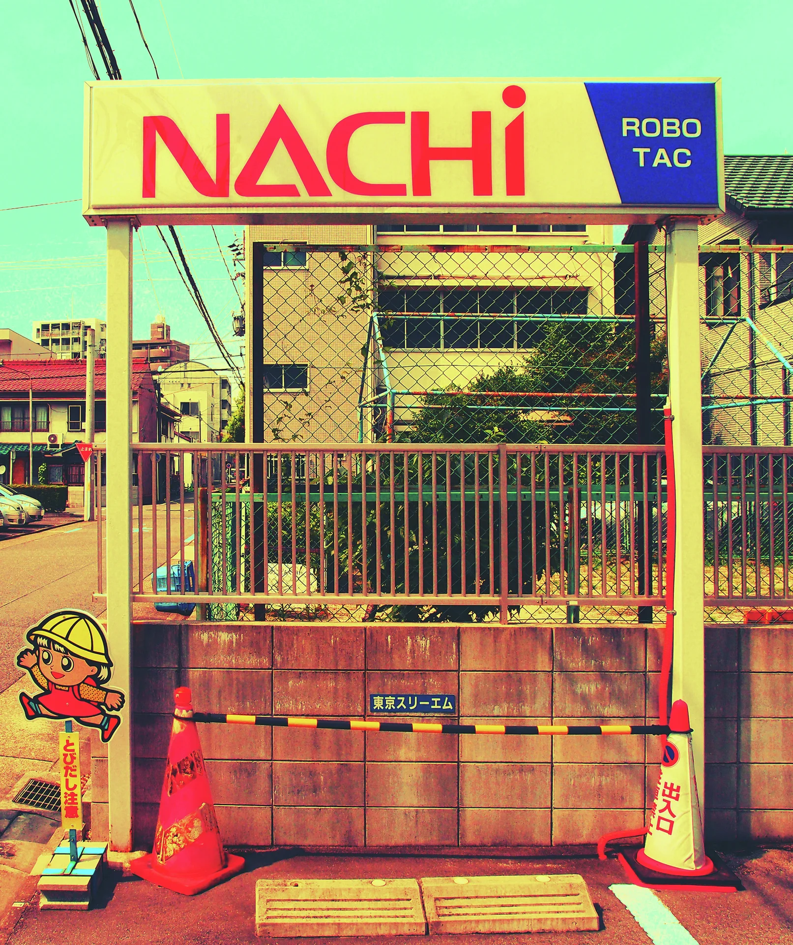A yellow Nachi sign crowns a wire-fenced industrial entrance bordered by orange cones and cartoon safety figures.