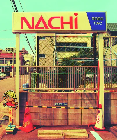 A yellow Nachi sign crowns a wire-fenced industrial entrance bordered by orange cones and cartoon safety figures.