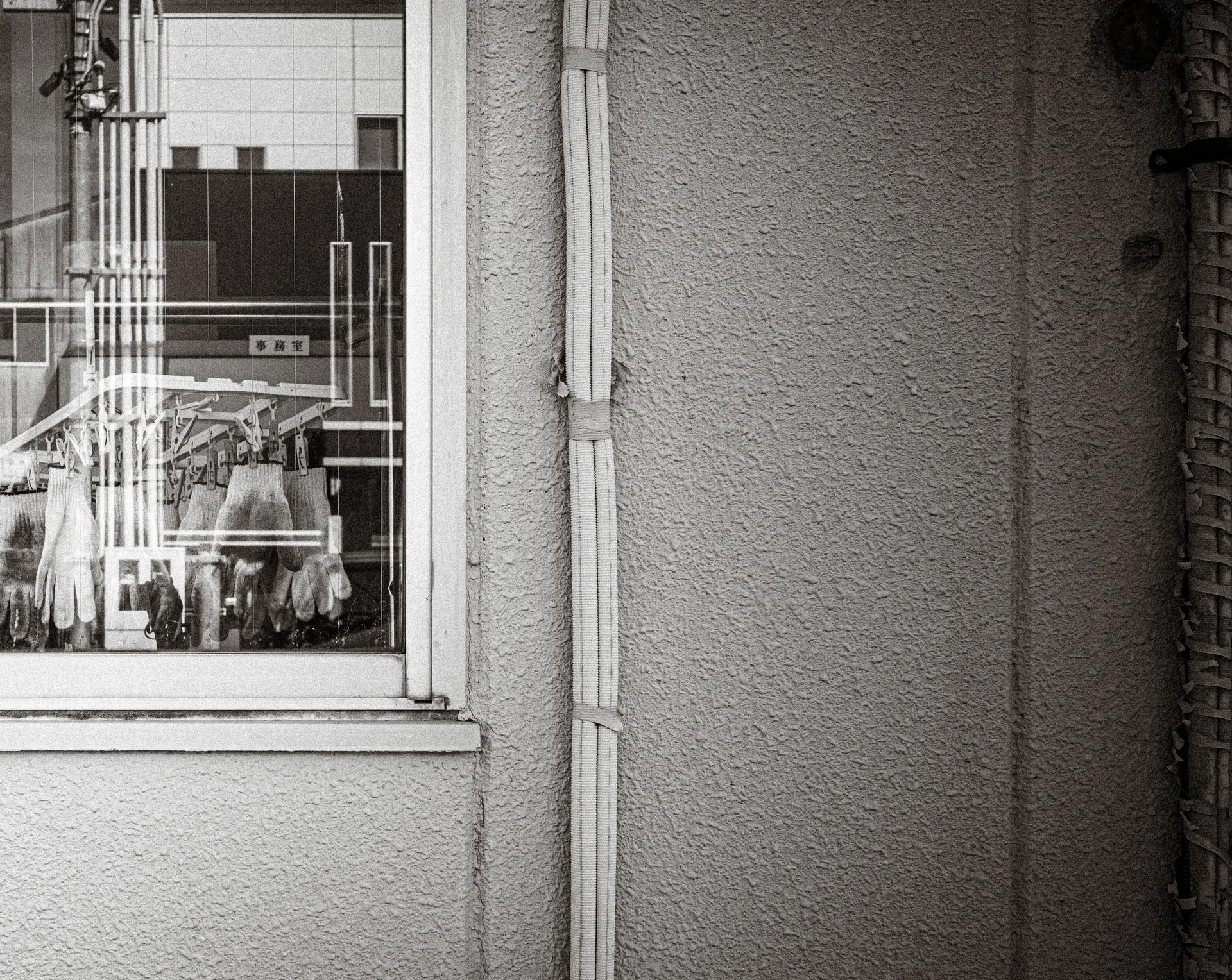 A geometric window grille reflects nearby buildings beside a textured wall and drainpipe in black and white.