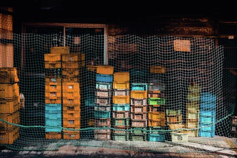 Stacks of turquoise, yellow, and pale crates stand behind green netting inside a dark storage opening.