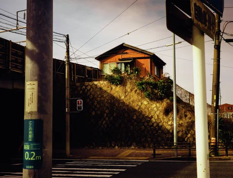 Sunlight illuminates a small wooden house above a stone retaining wall beyond foreground utility poles.