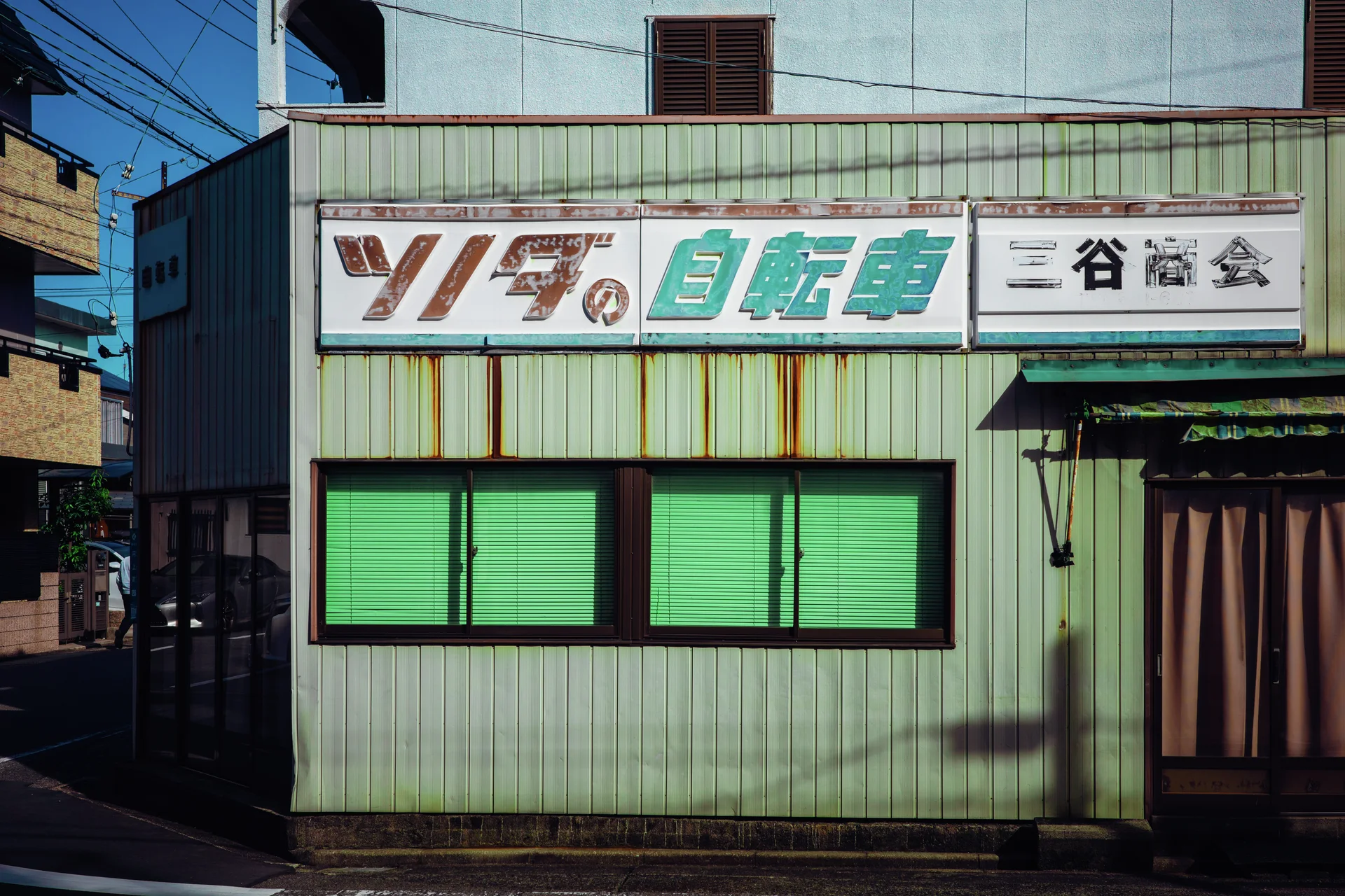 A rust-streaked corrugated storefront has green window blinds beneath a long sign and beside a brown entrance.