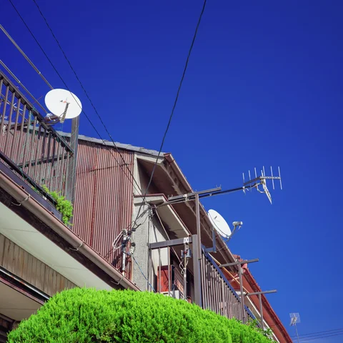 Satellite dishes and a television antenna project above corrugated rooftops beneath a deep blue sky.