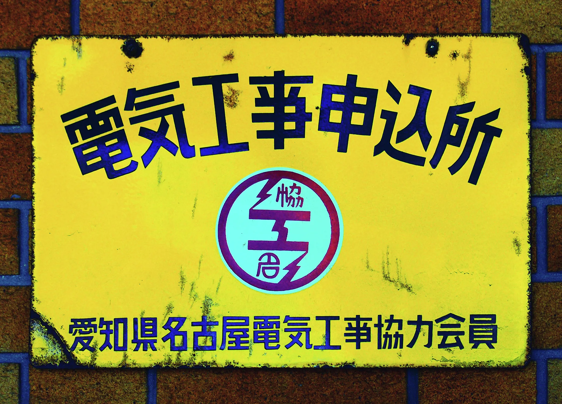 Dark blue Japanese lettering and a circular emblem fill a weathered yellow plaque mounted on brick.