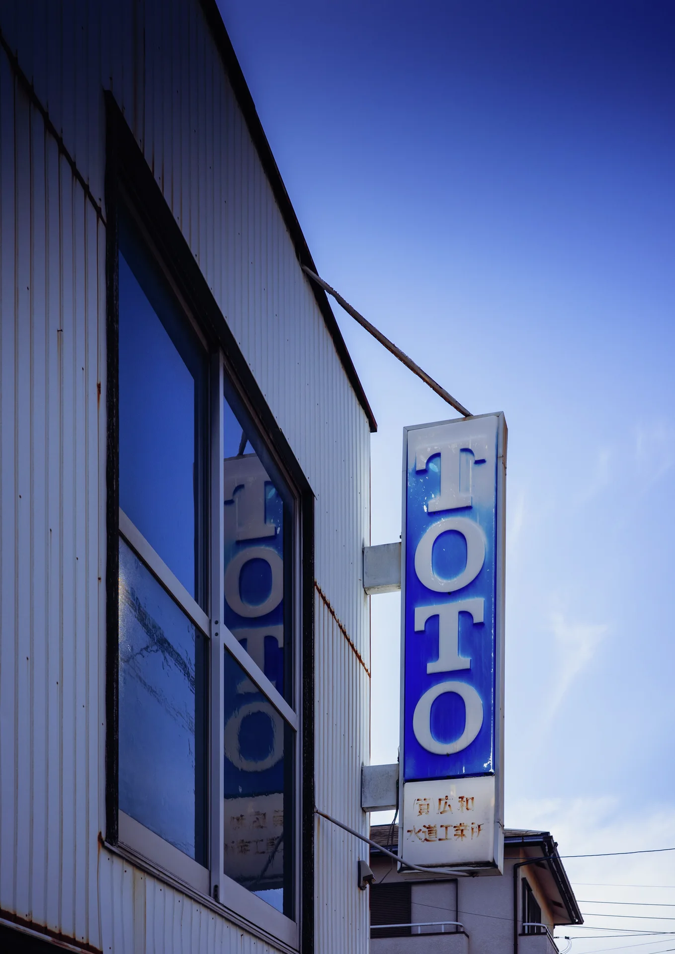 A blue vertical sign projects beside reflective windows against a clear blue sky.