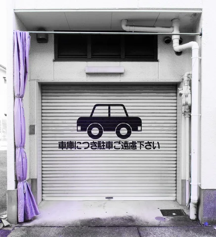 A black car silhouette and Japanese lettering decorate a closed pale garage shutter beneath exposed pipes.
