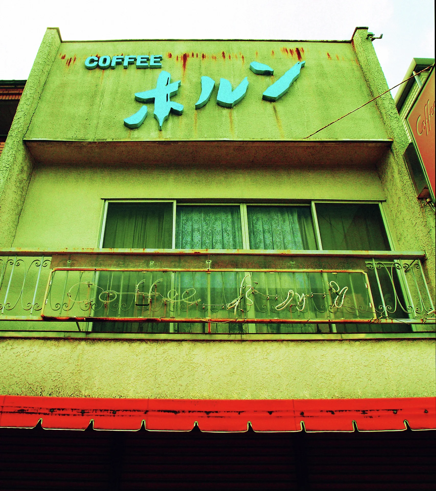 Turquoise raised lettering crowns a green coffee shop facade above a rusty balcony and red awning.