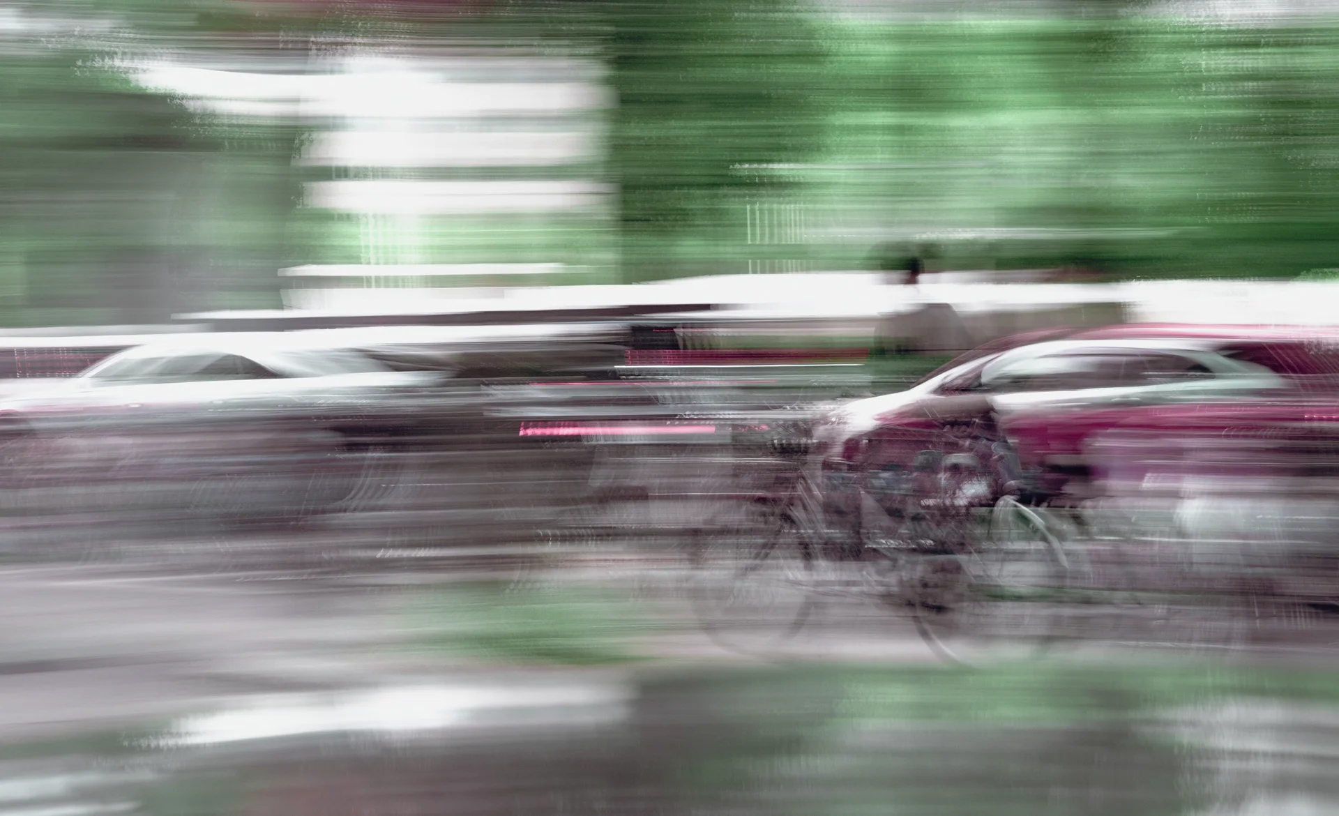 A blurred cyclist passes cars along a street with green and white horizontal motion streaks.