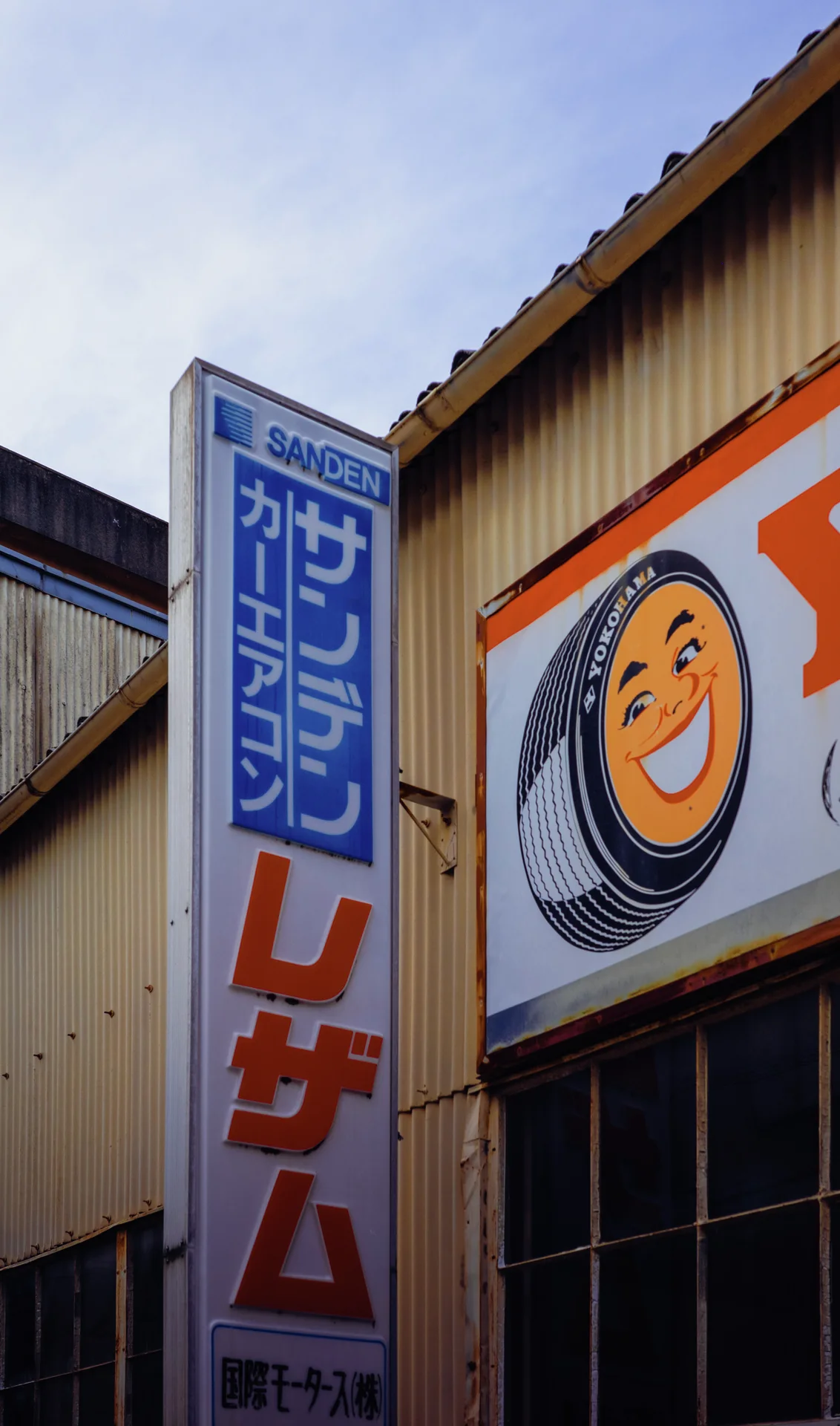 Vertical Japanese signage and a smiling circular emblem mark a corrugated building beneath a pale sky.