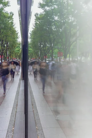 Pedestrians blur along a tree-lined sidewalk beside glass reflecting their figures and the pavement.