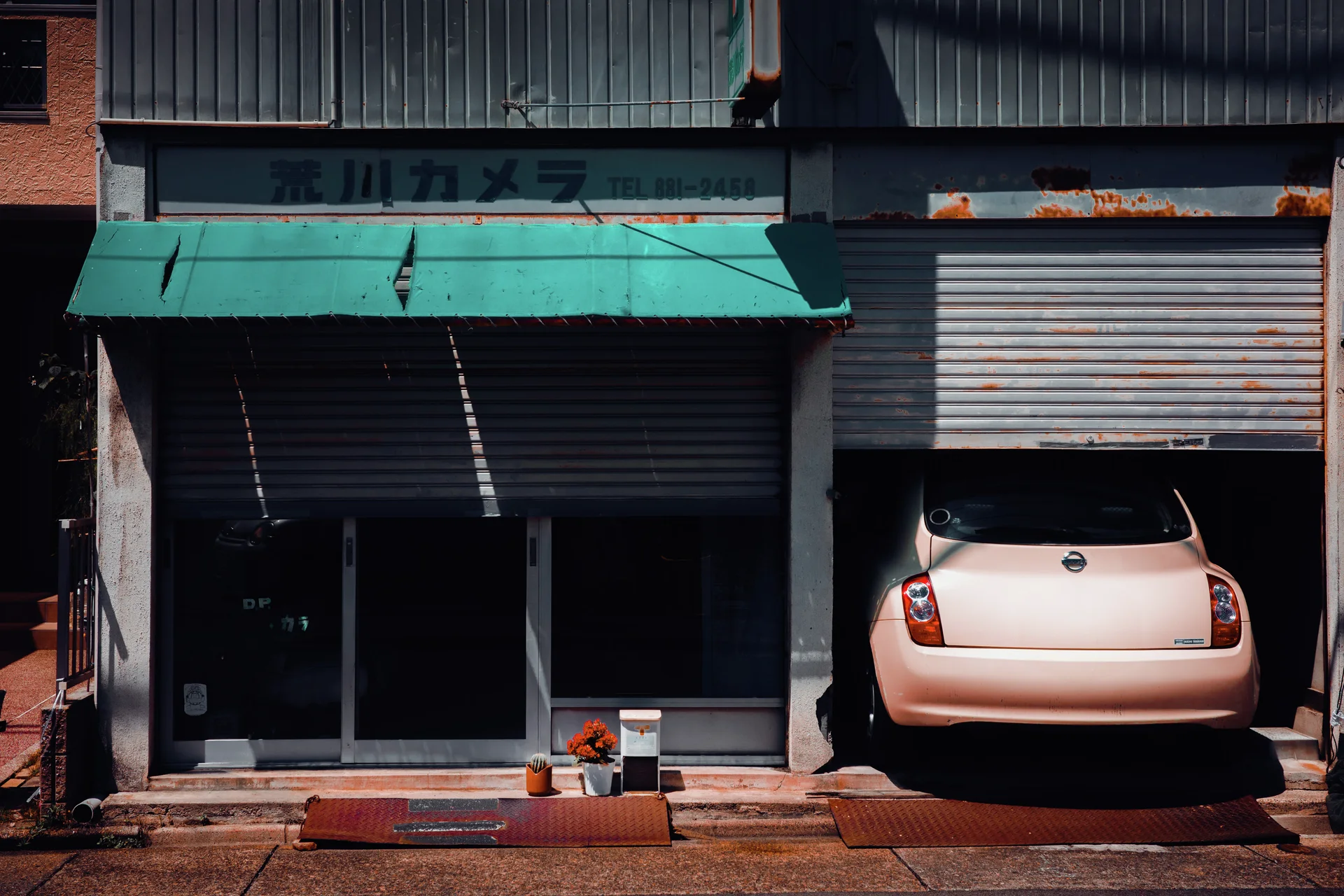 A pale pink car sits inside a partly shuttered garage beside a closed storefront with a turquoise awning.