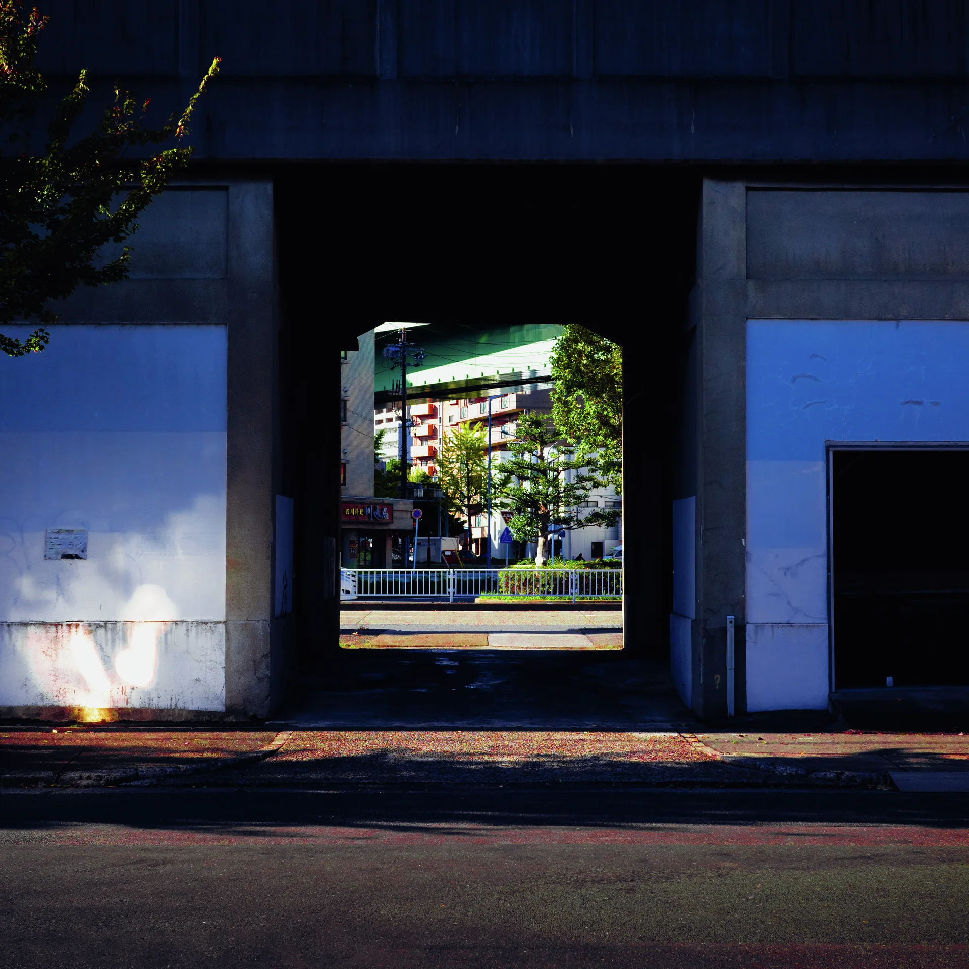 A dark passage through a blue industrial building frames a sunlit street and leafy trees beyond.