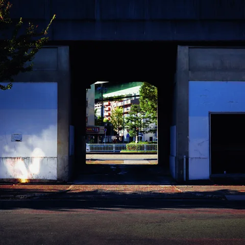 A dark passage through a blue industrial building frames a sunlit street and leafy trees beyond.