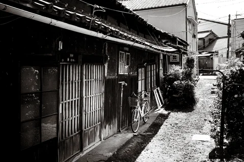 A bicycle leans against a weathered wooden house beside a sunlit gravel lane in monochrome.