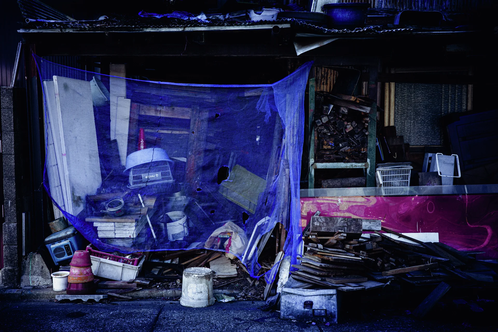 Blue mesh covers piled containers and equipment outside a cluttered weathered shed.