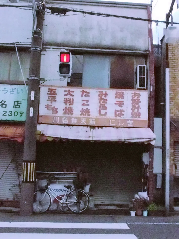 A bicycle rests before closed shop shutters beneath a faded menu sign and red traffic signal.