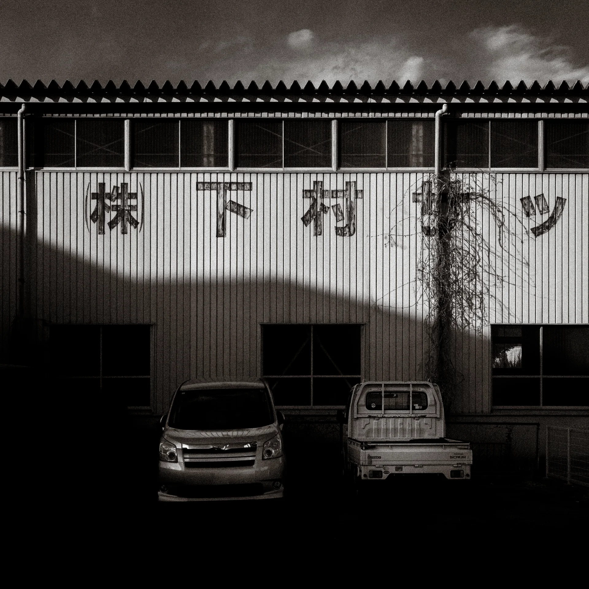 Two vehicles stand outside a corrugated warehouse crossed by broad shadows in black and white.