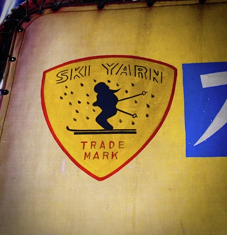 A black skiing figure decorates a yellow shield-shaped SKI YARN emblem on a stained pale wall.
