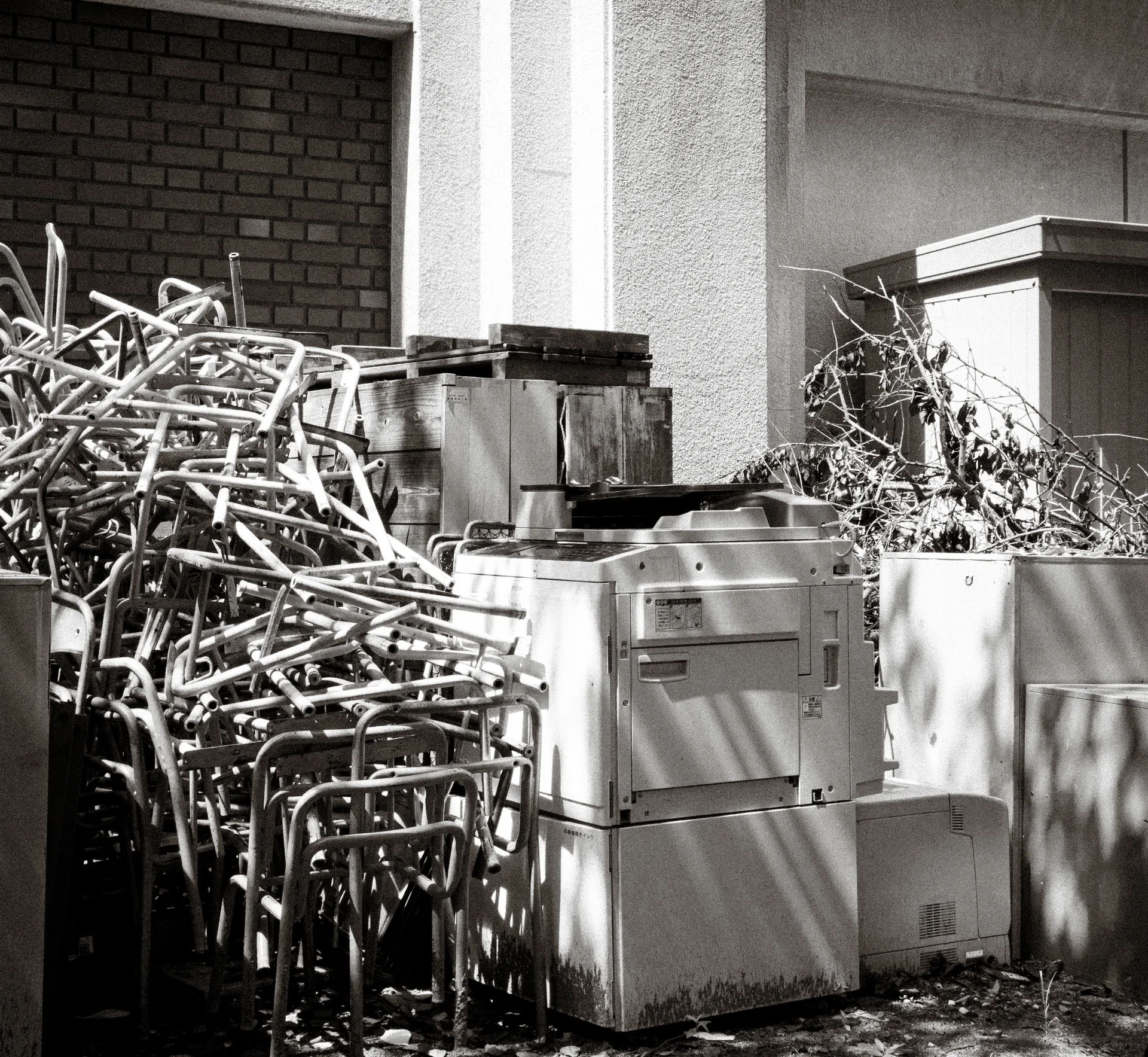 Stacks of metal frames and discarded appliances crowd a sunlit building corner in a black and white view.