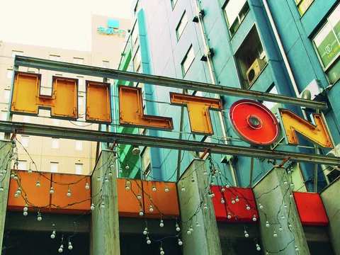 Large yellow and red Hilton letters hang across a metal frame above strings of lights between buildings.