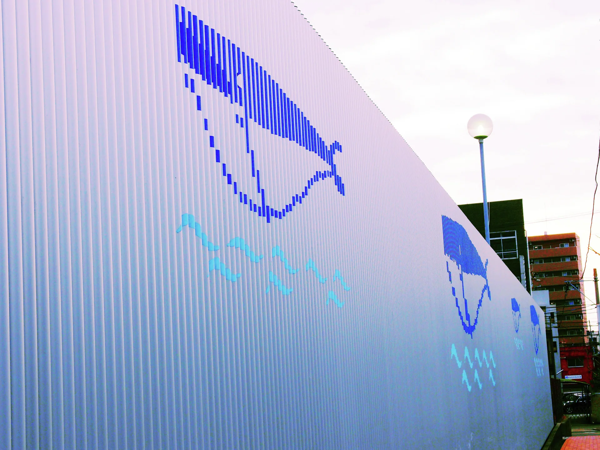 Blue stylized fish and pale wave marks decorate a long white corrugated wall beside a narrow walkway.